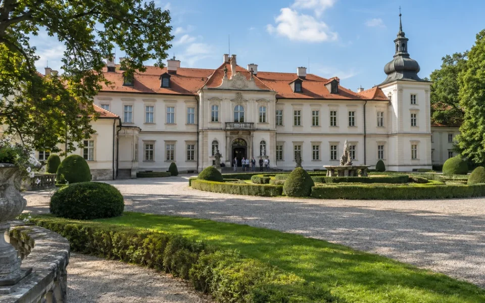 Zámek s upraveným parkem za teplého pozdního léta, na nádvoří několik návštěvníků čeká na prohlídku
