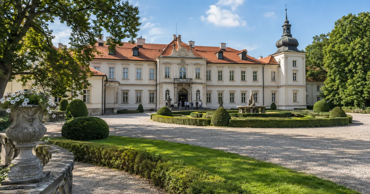 Zámek s upraveným parkem za teplého pozdního léta, na nádvoří několik návštěvníků čeká na prohlídku